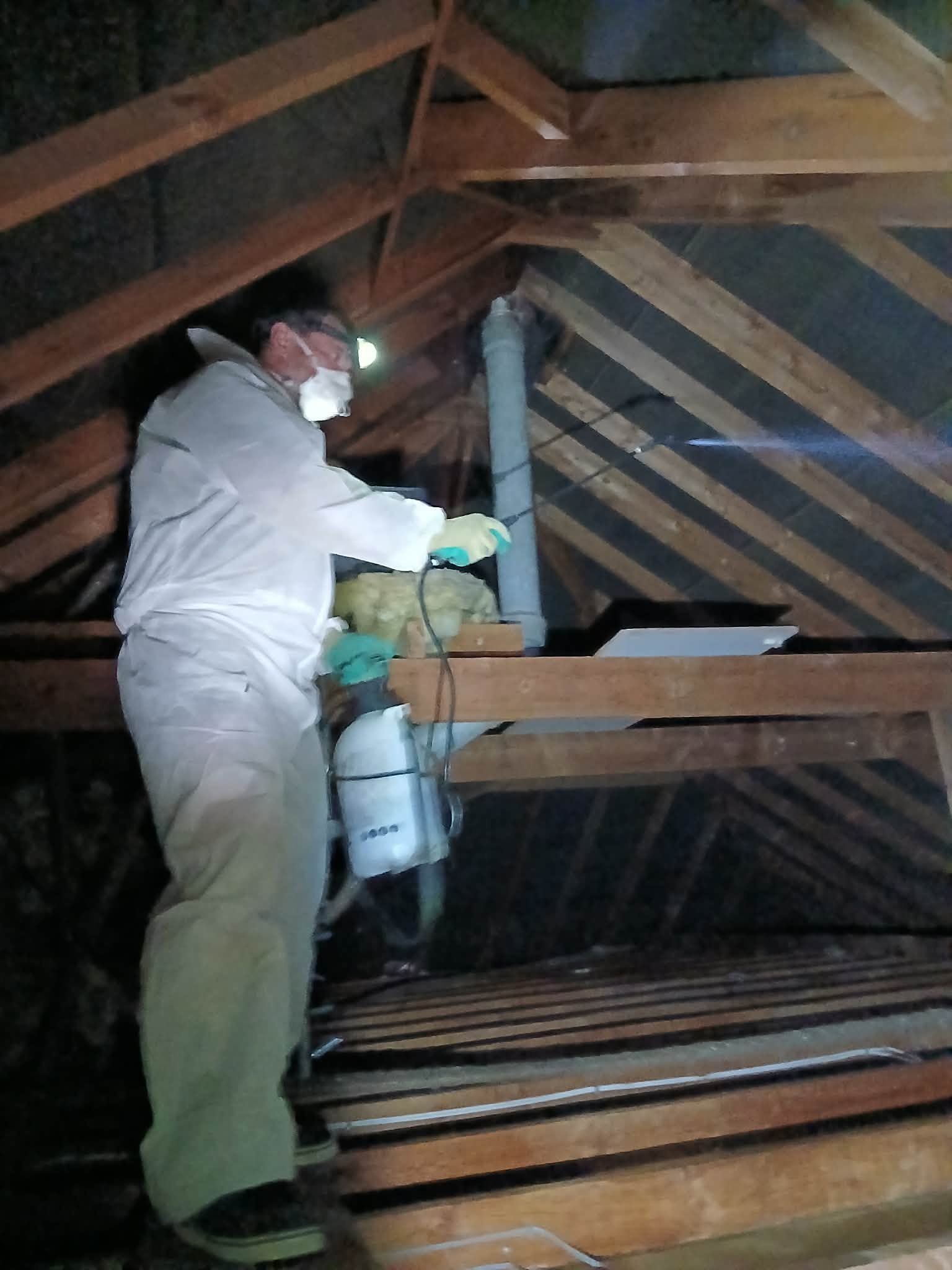 Worker in protective clothing and respirator working among attic rafters, insulation, ducts, and wooden beams.